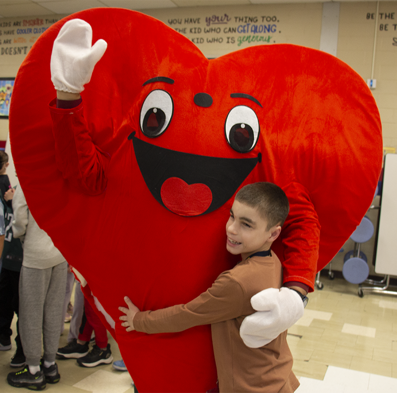 A person in a large red heart costume waves a hand and gets a hug from a little boy.