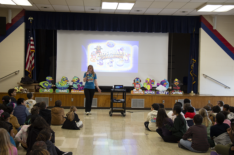 A woman stands in front of a large group of kids. Behind her is a graphic about heart health.