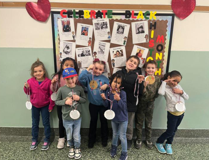 A group of young elementary students wear their paper gold medals and stand in front of a bulletin board for Black History Month.