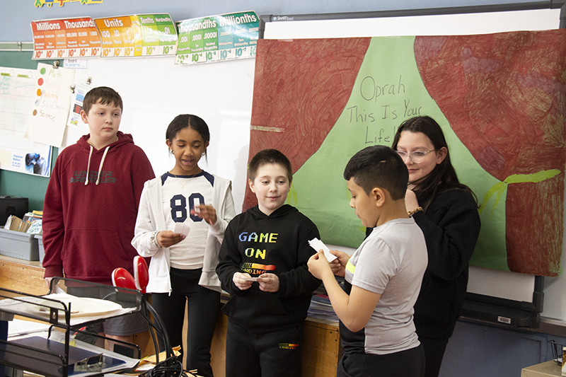 A group of fifth-grade students stand in the front of a room and make a presentation. Behind them is a sign that says This is your life Oprah Winfrey.