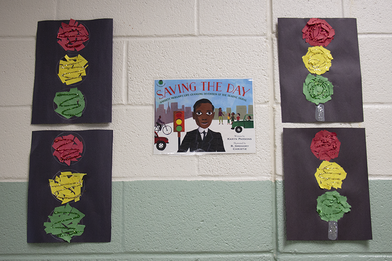 Stop lights made from construction paper hang on a wall. In the center is a picture of a man who invented them.