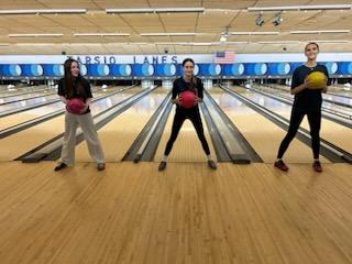 Three high school kids stand in front of bowling lanes with bowling balls.