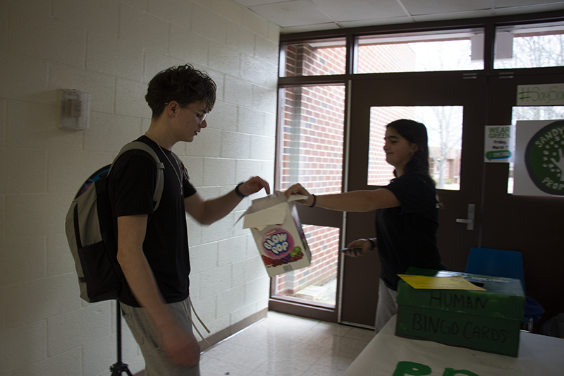 Two high school kids. One is offering a plastic big of lollipops and the other is picking one out.