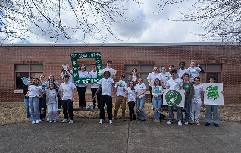 A group of about a dozen high school kids, all wearing green and white t shirts. they are holding signs that say "Say Something" and "Sandy Hook Promise."