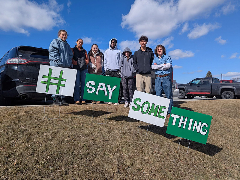 A group of seven high school kids outside on a slope. They have yard signs that say #SaySomething.