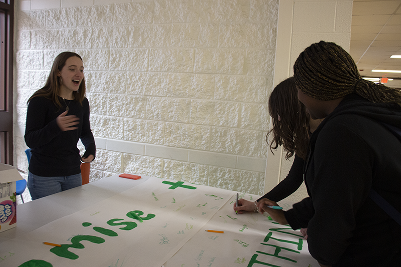 Two high school kids lean over a large white banner that says Say Something. They are signing it.