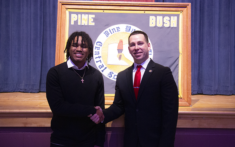 A high school junior on the left shakes hands with a man dressed in a suit. Behind them is a framed banner that says Pine Bush high School.