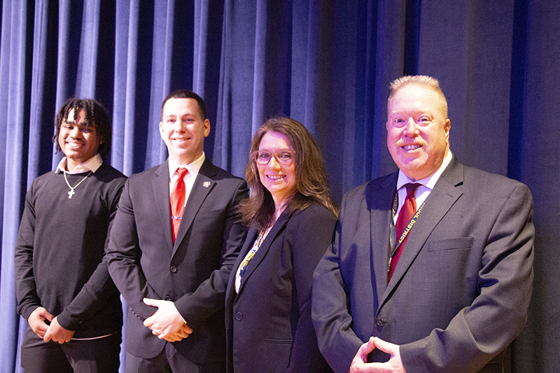 A high school student on the left next to three adults. All are smiling.