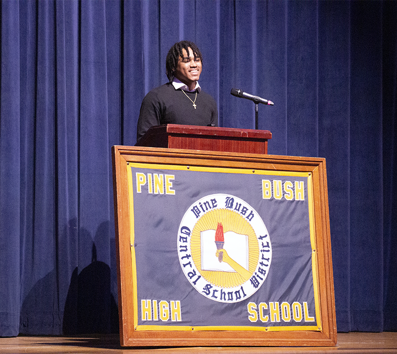 A young man smiles as he speaks from a podium. In front of it is a large framed banner that says Pine Bush High School.