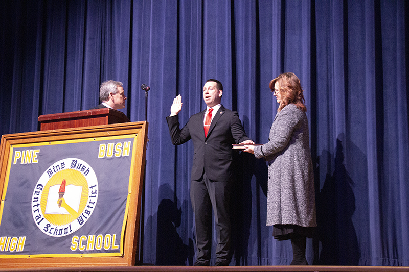 A man in the center holds up his right hand. Another man on the left talks and a woman on the right is holding a bible.